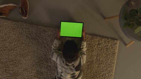 Fotografía-Cenital-De-Un-Niño-Acostado-Sobre-Una-Alfombra-En-Casa-Jugando-Juegos-O-Transmitiendo-A-Una-Tableta-Digital-De-Pantalla-Verde-Por-La-Noche-1